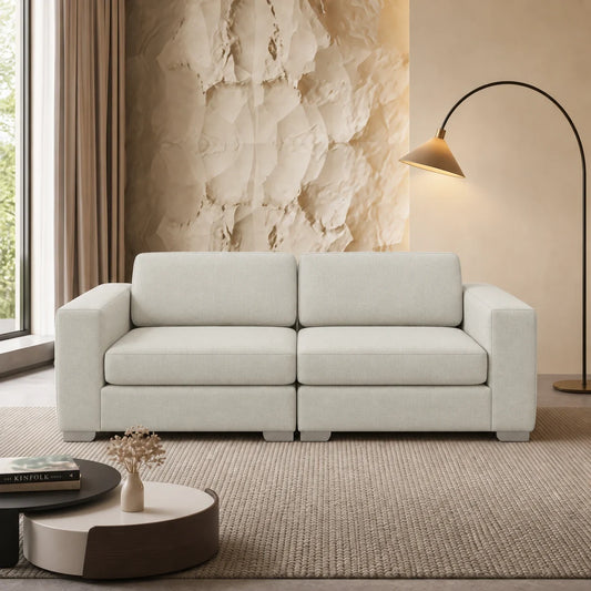 A modern two-seater sofa in a light beige fabric, styled in a contemporary living room setting with a textured rug, a sculptural beige wall, a black floor lamp, and a round coffee table.